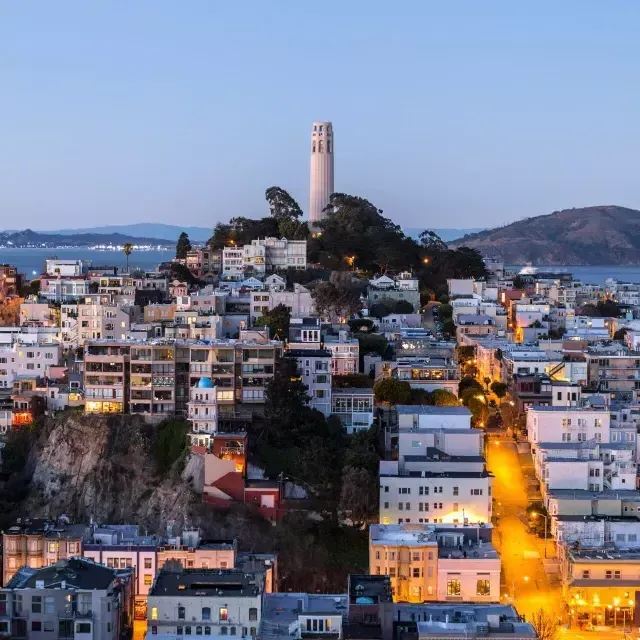 Coit Tower de San Francisco al anochecer, con calles iluminadas frente a ella y la Bahía de San Francisco detrás.